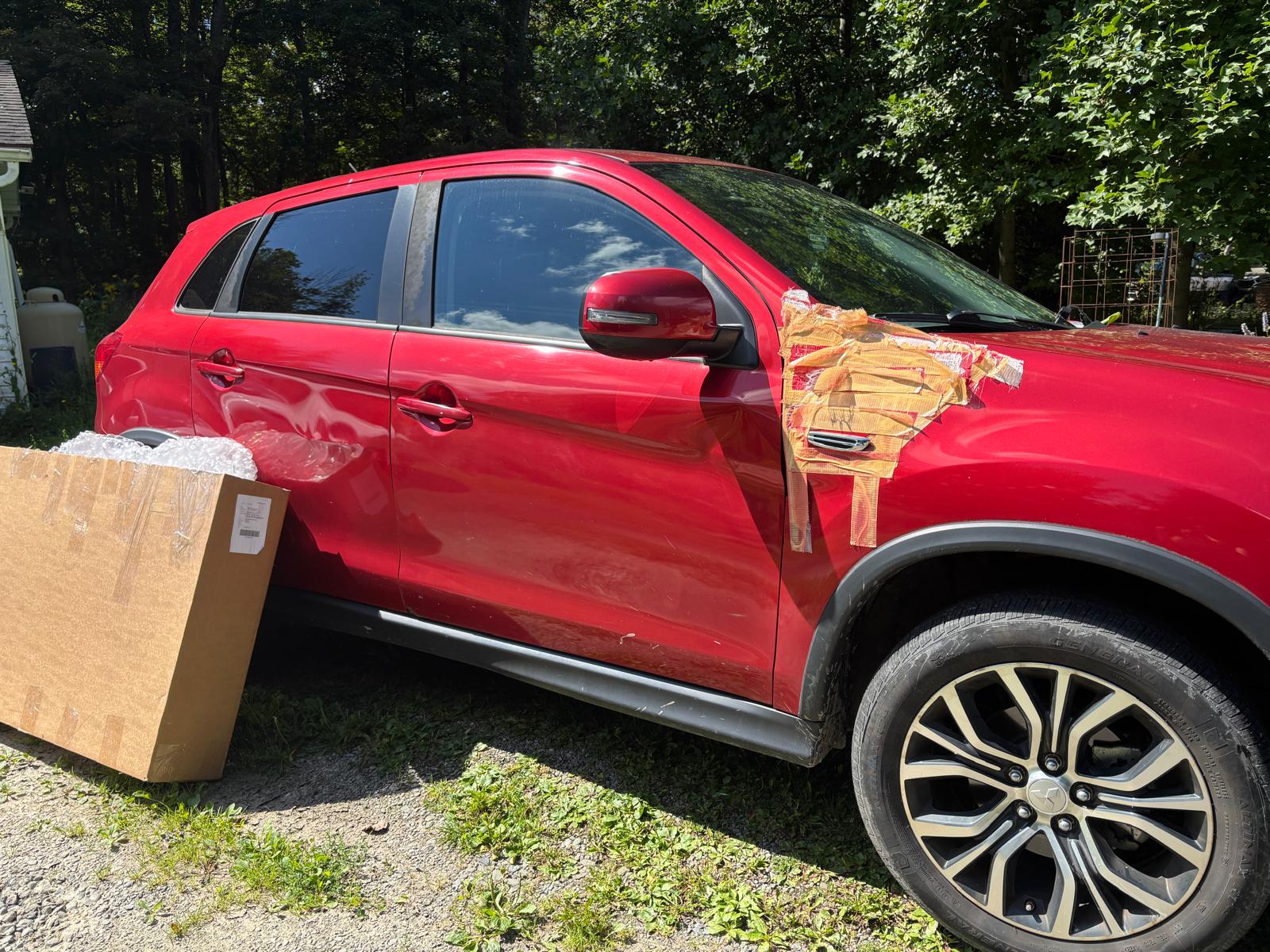 Red Mitsubishi Outlander before exterior part replacement with a boxed part beside it